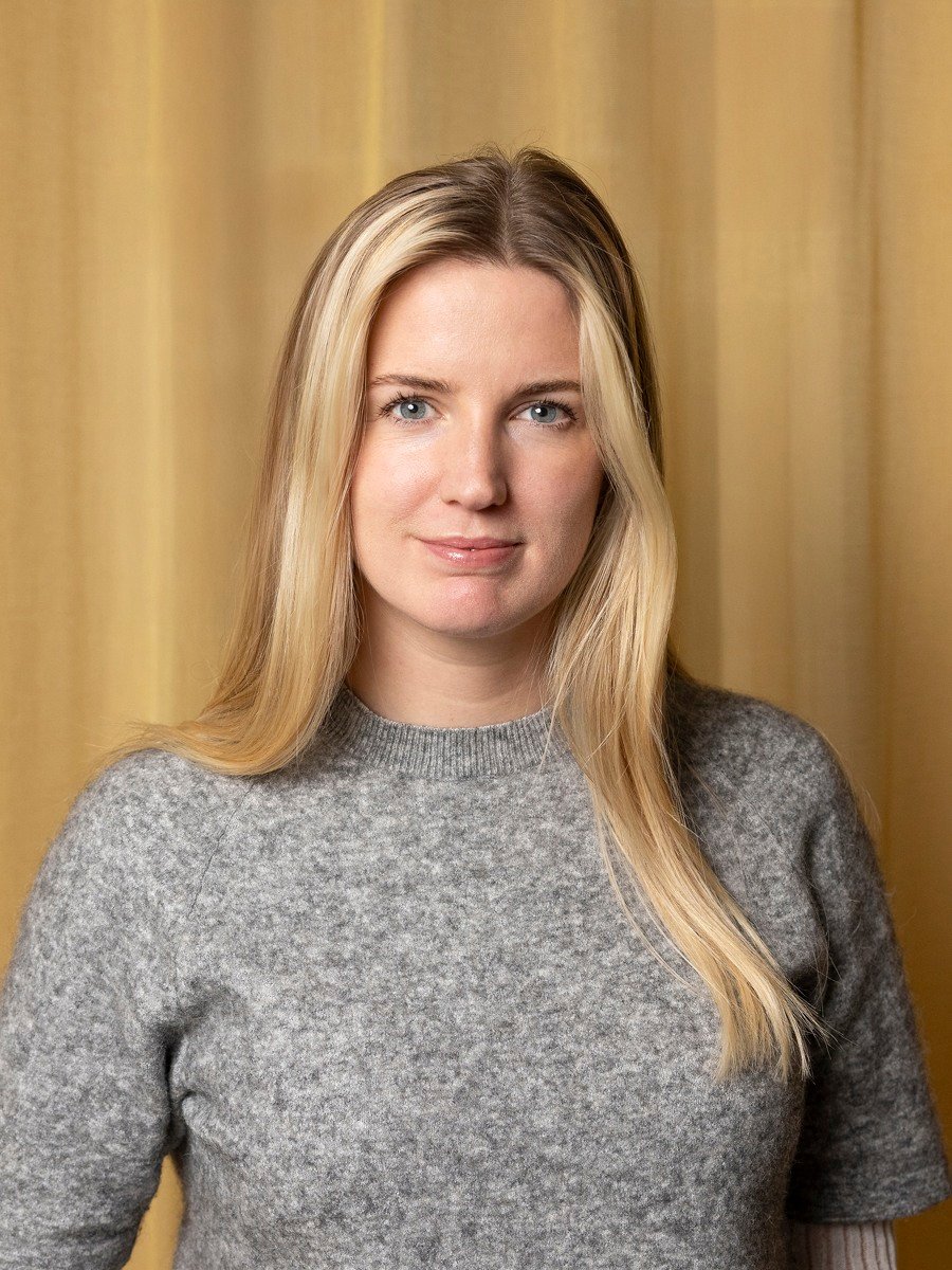 En ung blond kvinna står framför en gul gardin.