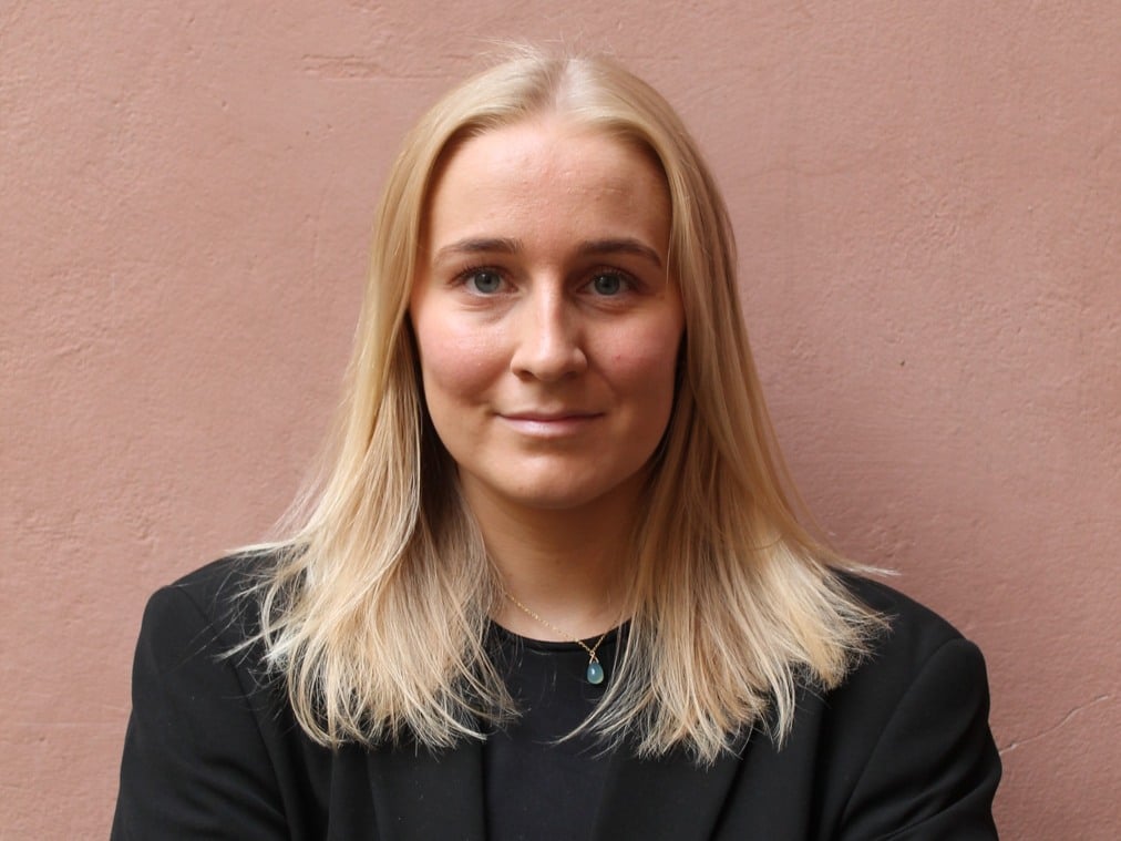 En kvinna med blont hår står framför en rosa vägg.