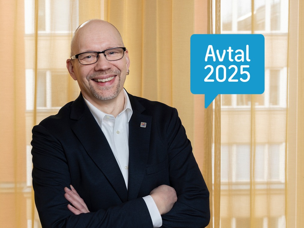En glad person i kostym och glasögon står med armarna i kors.